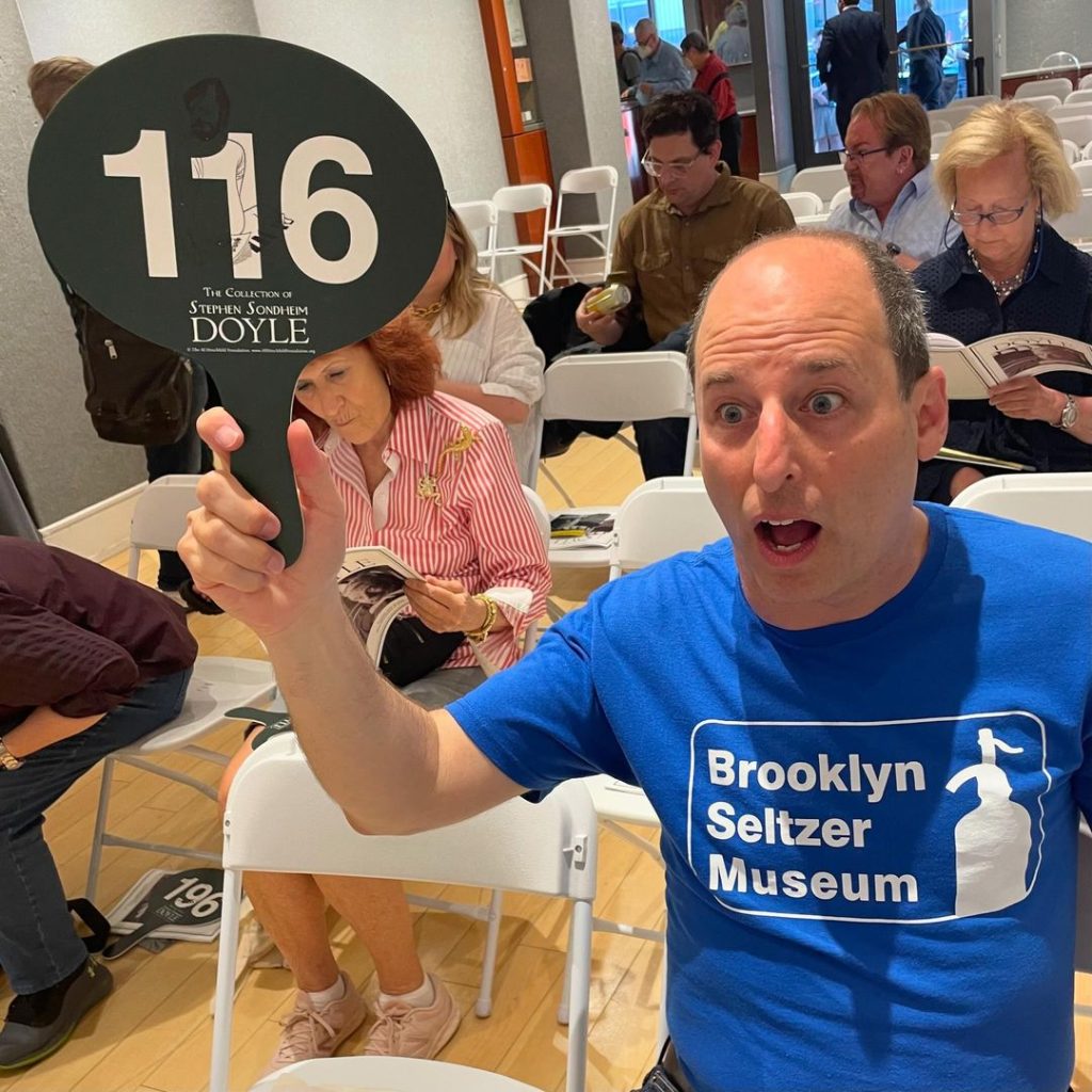 A man eagerly holds up an auction paddle with a large number 116 on it